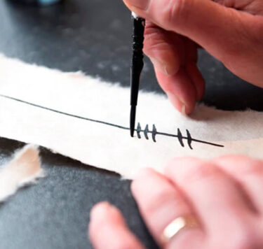 Black background with someone writing Ogham inscriptions onto a piece of paper