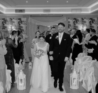 Couple walks down aisle in elegant, warmly-lit hotel wedding venue with floral decorations and applauding guests.