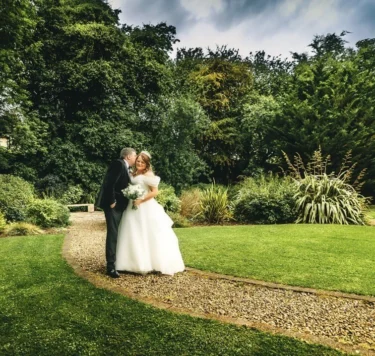 Bride and groom embrace on gravel path in lush hotel gardens with manicured lawn and mature trees, ideal for weddings
