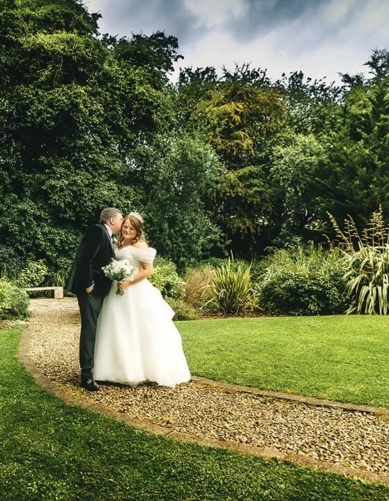 Bride and groom embrace on gravel path in lush hotel gardens with manicured lawn and mature trees, ideal for weddings